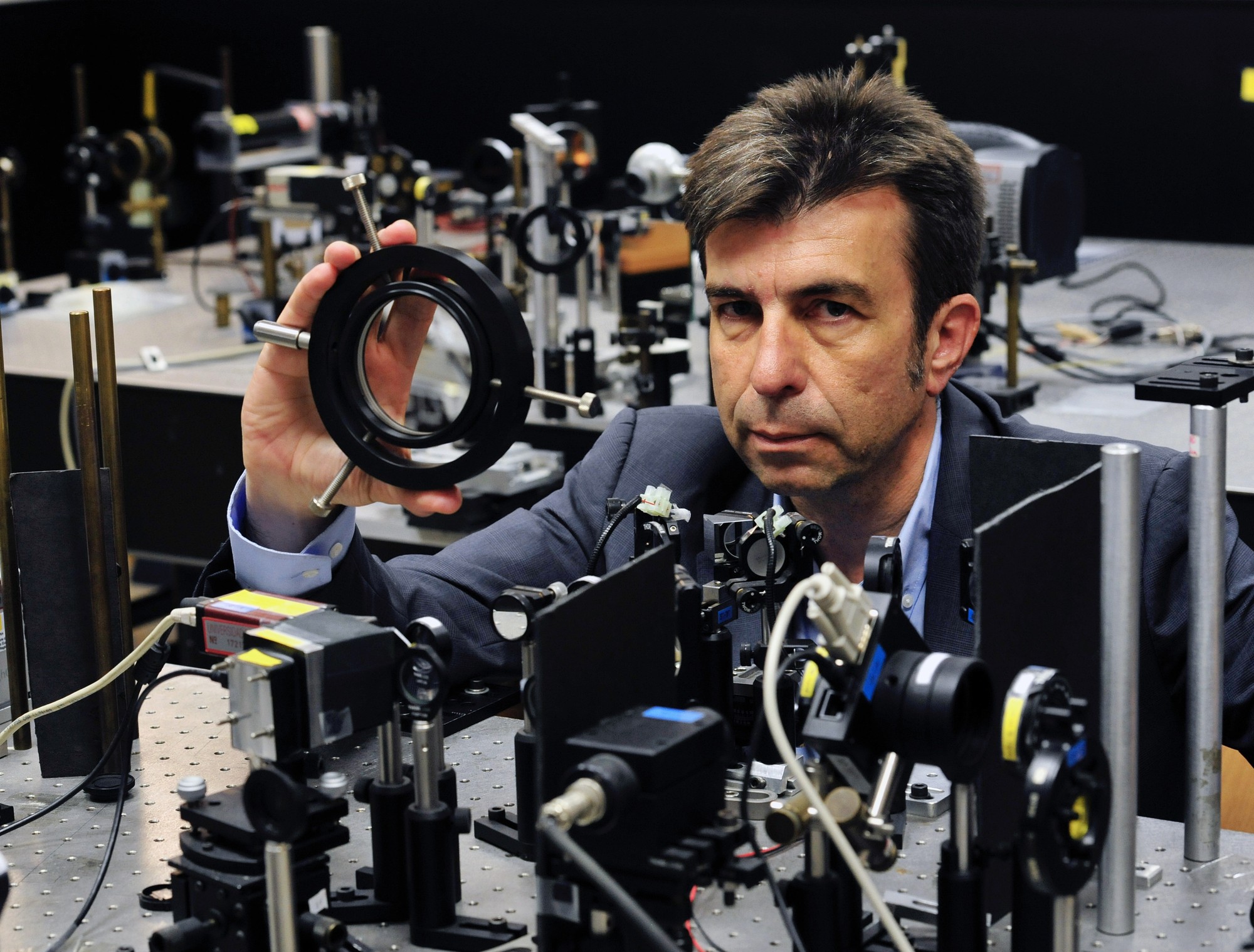 Pablo Artal holding a mounted lens, surrounded by optical instruments in the laboratory.